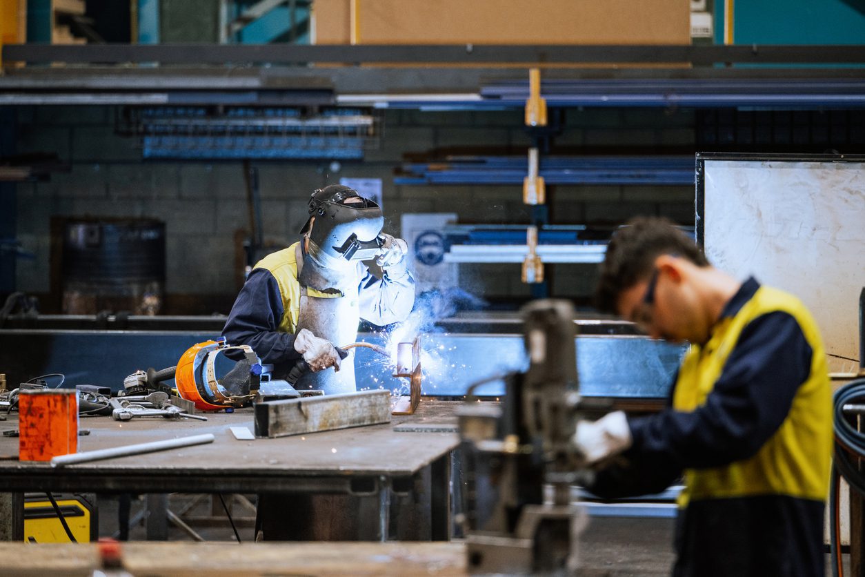 Diverse workers in protective gear welding metal in an industrial factory setting with sparks flying around brightly illuminating the area