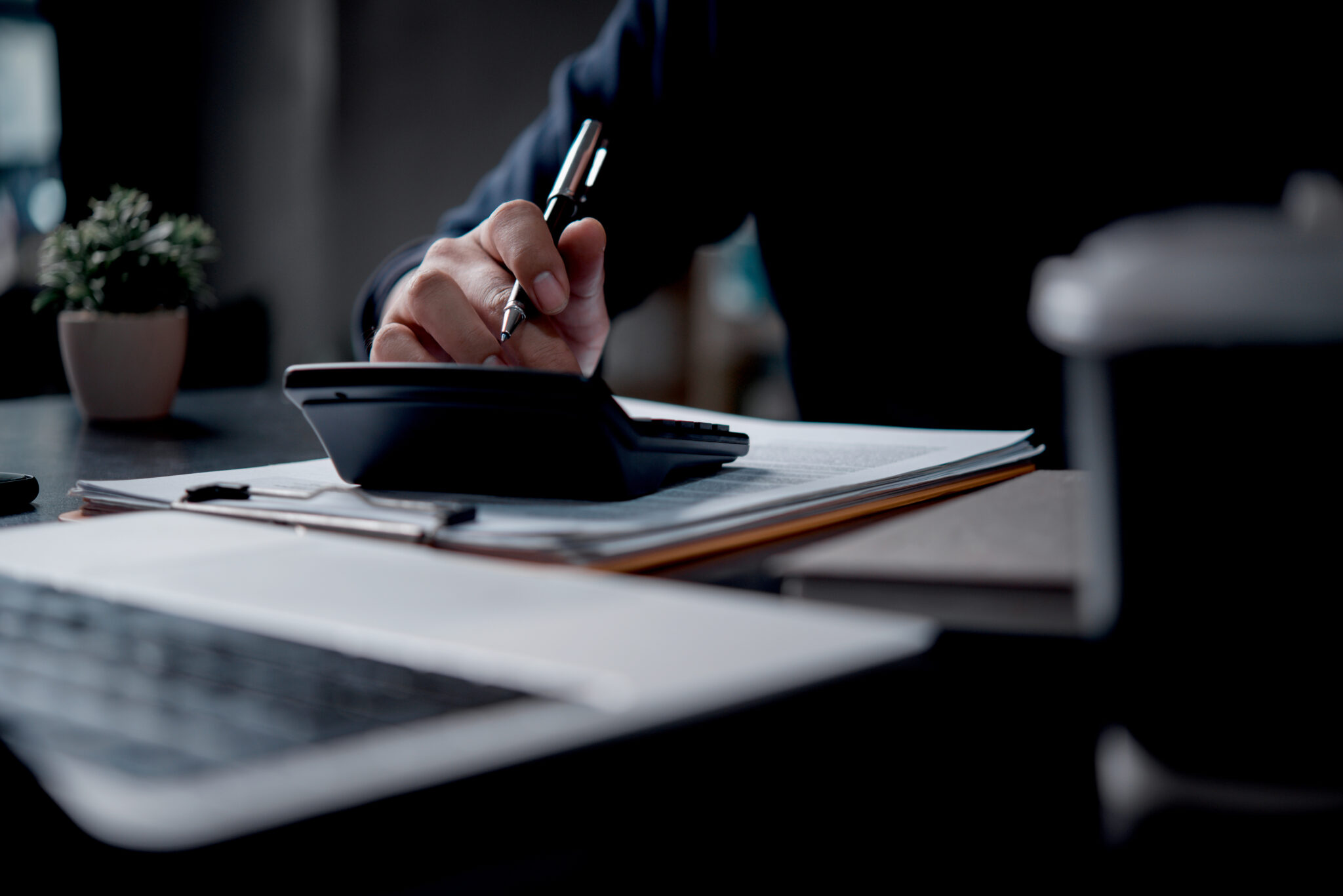 A business professional using a calculator and pen to review financial documents in an office setting. The scene symbolizes financial planning and business management tasks.